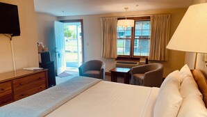 Quarto de hotel com cama, escrivaninha, duas cadeiras, televisão e janela com cortinas.