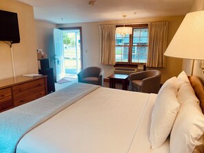 Une chambre d’hôtel comprenant un lit, un bureau, deux chaises, une télévision et une fenêtre avec des rideaux.