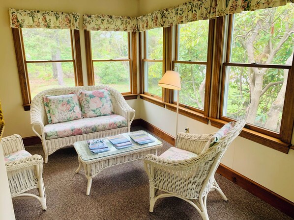 A cozy room with a wicker sofa, two chairs, a small table, and large windows with a view of trees. - The Chatham Motel (Chatham)