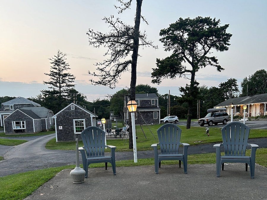 A paved outdoor area with three Adirondack chairs, a street lamp, and a row of houses in the background.