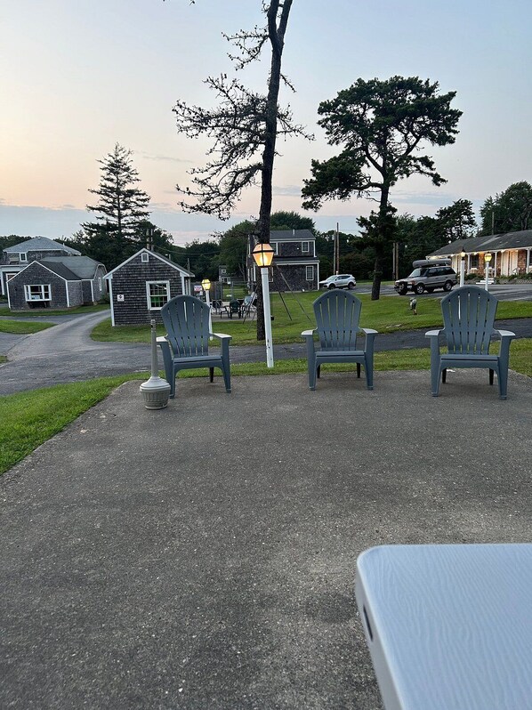 Un espace extérieur aménagé avec une allée pavée, trois chaises Adirondack, un lampadaire et une rangée de maisons en arrière-plan.