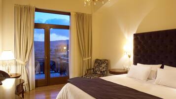 Quarto de hotel com uma cama grande, uma cadeira, uma mesa e vista para uma paisagem montanhosa através de uma porta de vidro.