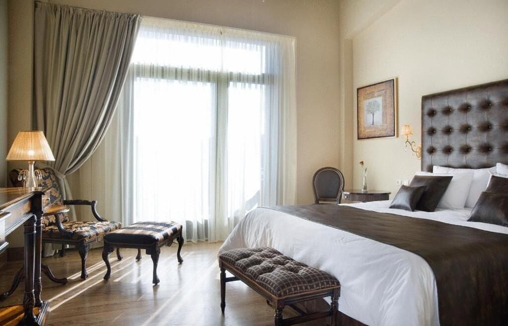 A hotel room with a large bed, a desk with a lamp, a chair, and a window with curtains.