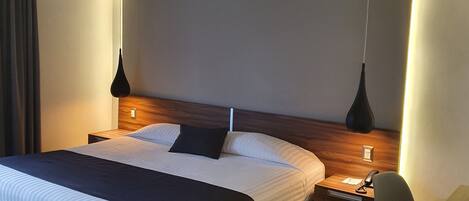 A modern hotel room with a large bed, a wooden nightstand, a desk, and a hanging lamp.