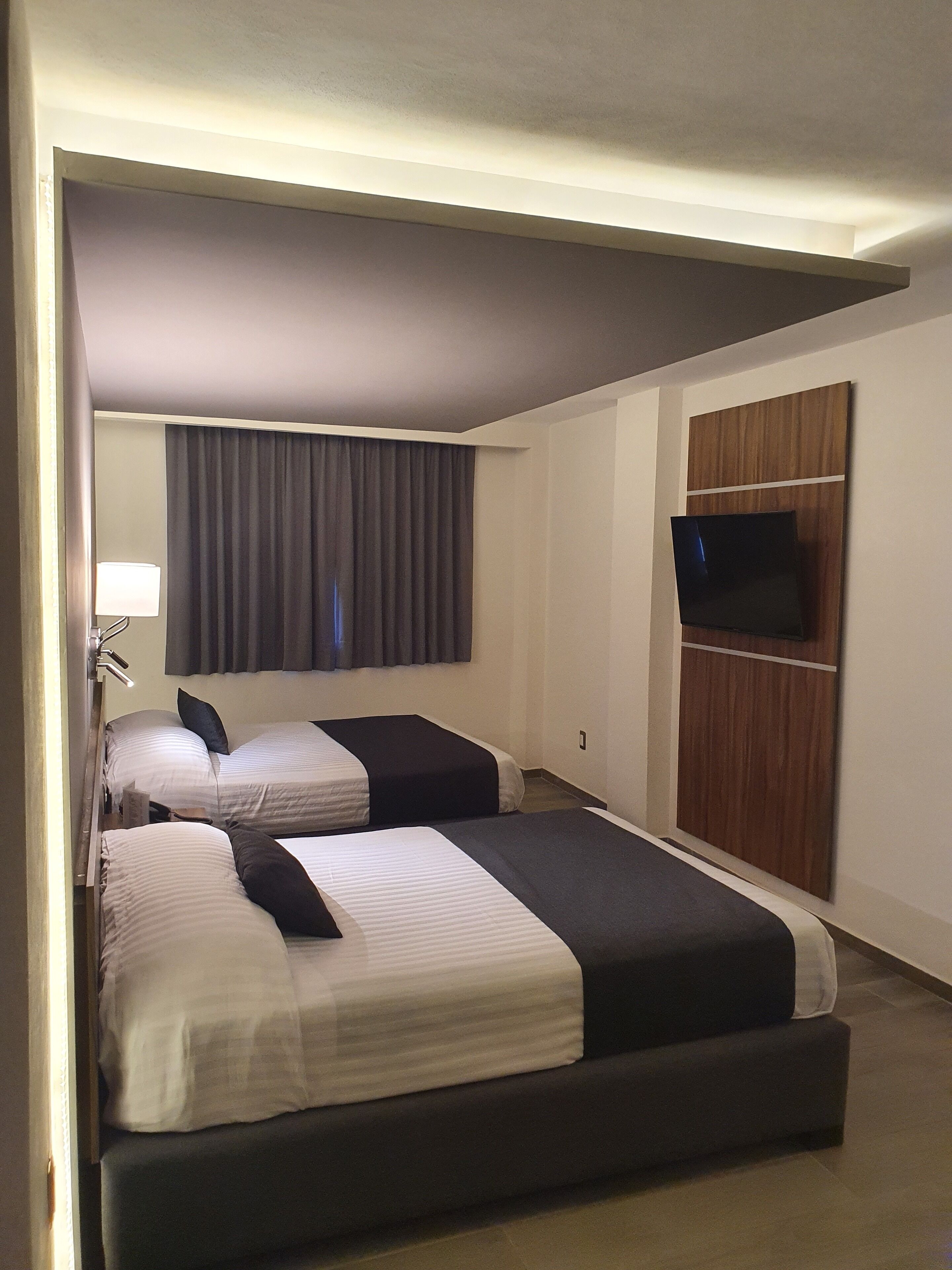 Habitación de hotel con dos camas, un televisor montado en un panel de madera, una lámpara y un ventanal con cortinas.