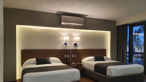 A hotel room with two beds, a wooden headboard, wall-mounted air conditioning, bedside lamps, and a view through a window. - Estanza Hotel & Suites (Morelia)