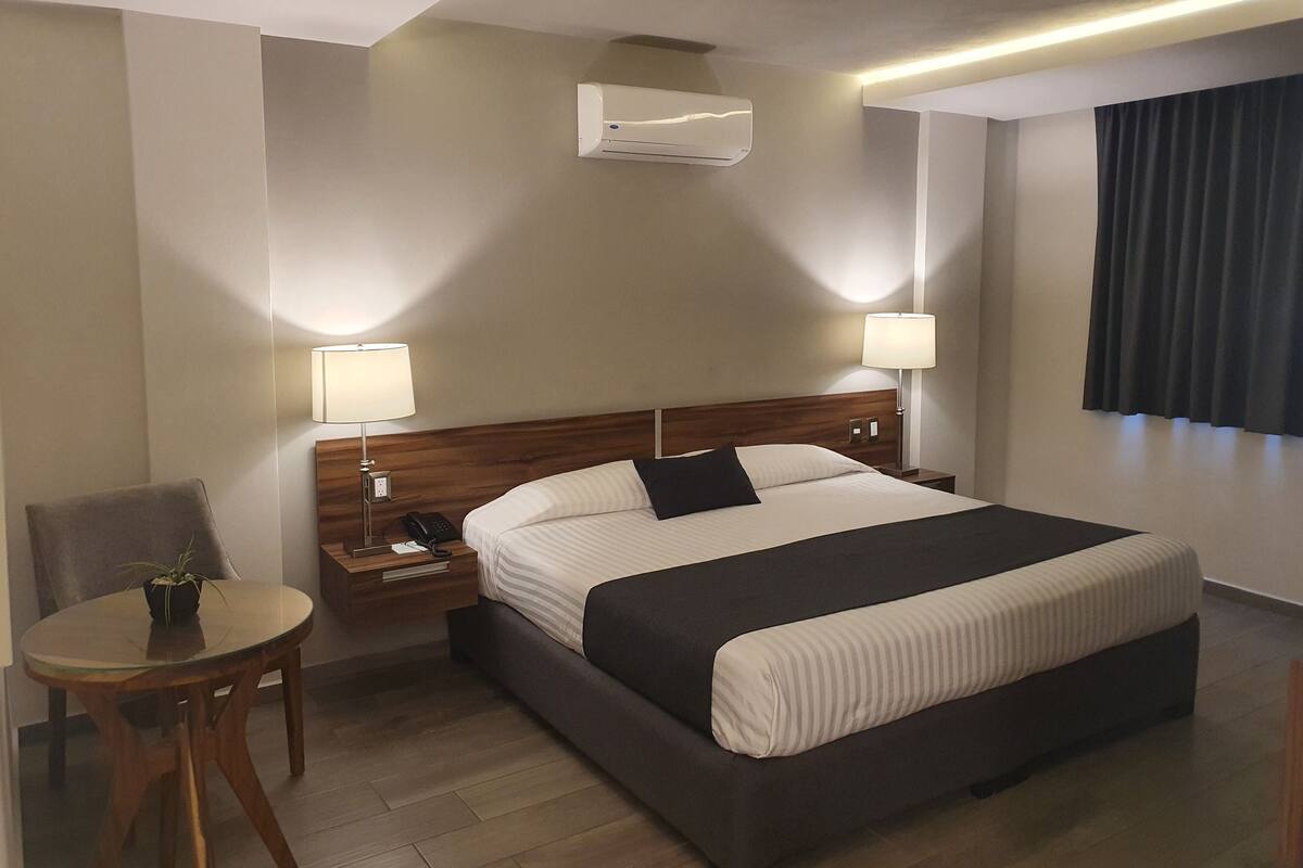 Ein Hotelzimmer mit Bett, Nachttischen, einer Lampe, einem Sessel und einer Klimaanlage.