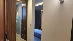 A hotel corridor with a wooden door, a wall-mounted light fixture, and a bed in the background. - Estanza Hotel & Suites (Morelia)
