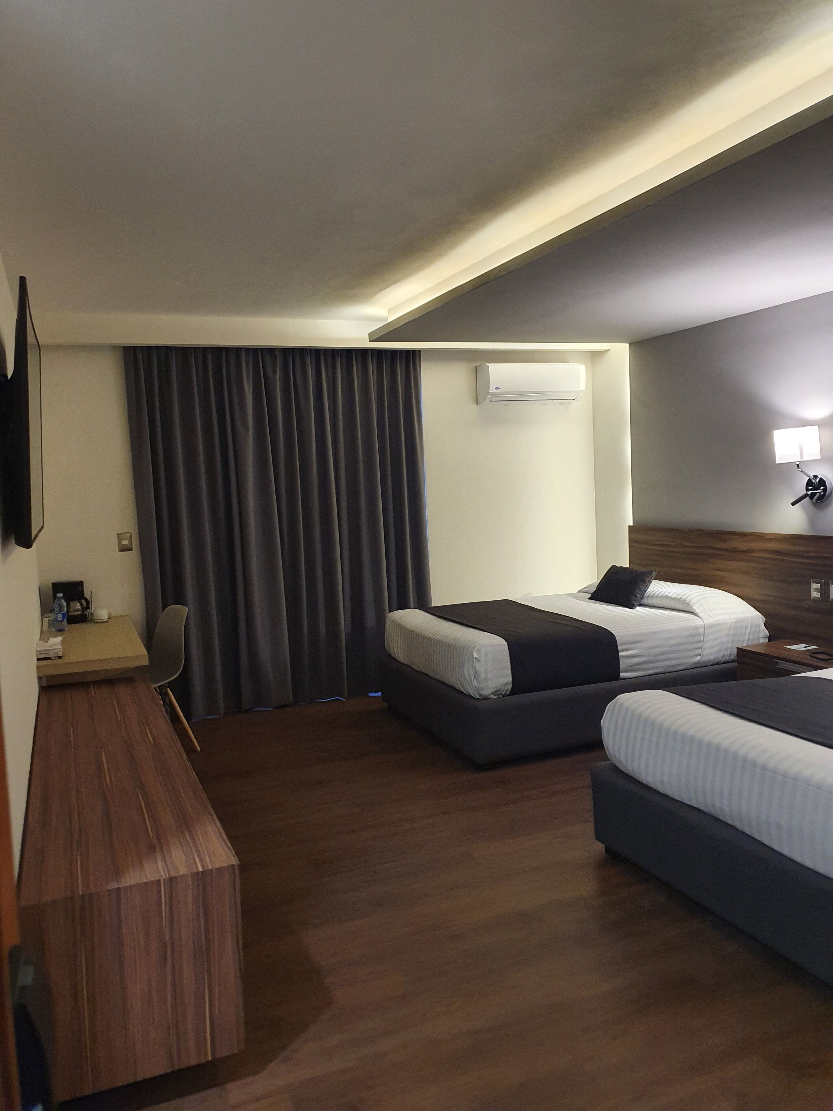 Habitación de hotel con dos camas, un escritorio, una silla, un equipo de aire acondicionado y una lámpara de pared.