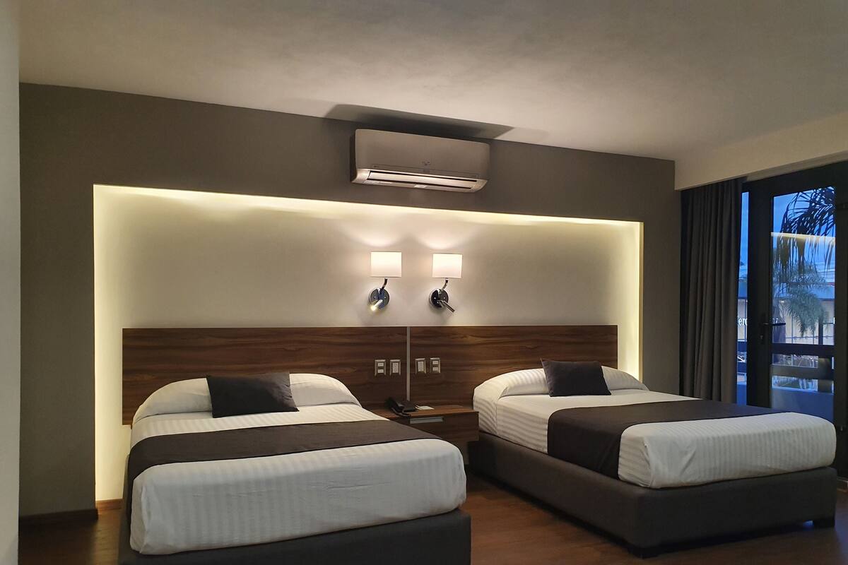 Ein Hotelzimmer mit zwei Betten, einem Holz-Kopfteil, einer Wand-Klimaanlage, Nachttischlampen und Blick ins Freie.