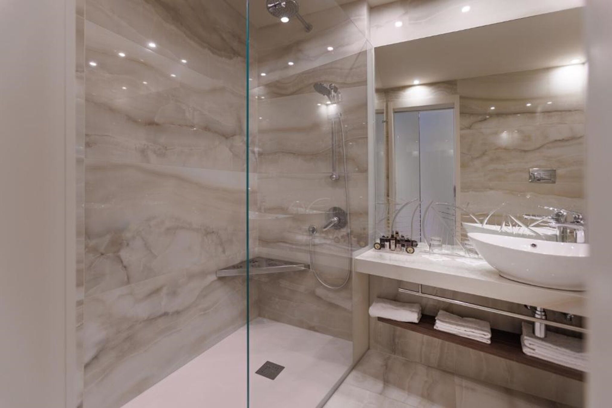 Un baño moderno con ducha envuelta en vidrio, paredes de mármol y un espejo grande sobre un lavabo blanco con pileta.