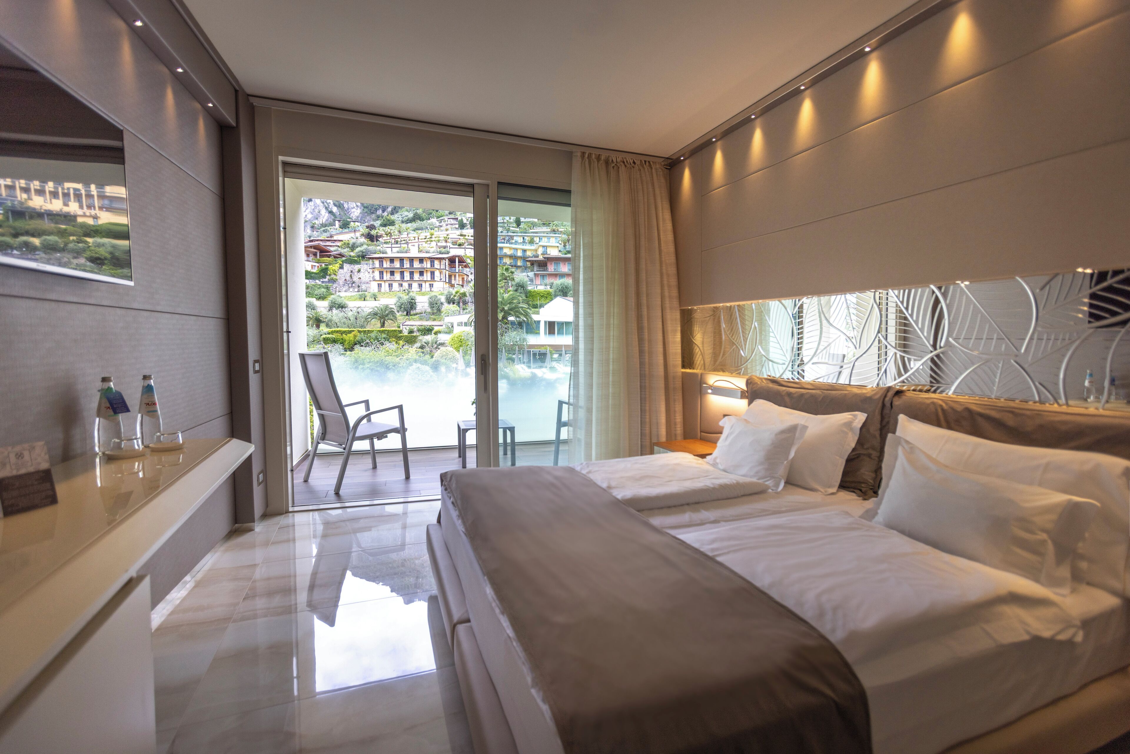 Ein modernes Hotelzimmer mit einem großen Bett, einer Glastür, die auf einen Balkon mit Blick auf eine Hügel landschaft führt, und einem Empfangstresen mit Wasserflaschen.
