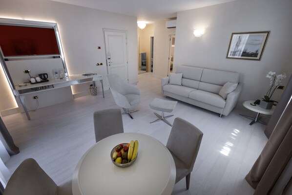 A modern living room with a white sofa, a dining table with fruit, and a kitchen area with a red appliance. - Park Hotel Imperial Centro Tao Natural Medical Spa (Limone sul Garda)