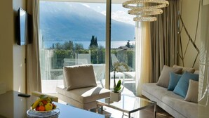A modern living room with a sofa, a coffee table, and a TV. There is a large window with a view of mountains and a lake. - Park Hotel Imperial Centro Tao Natural Medical Spa (Limone sul Garda)