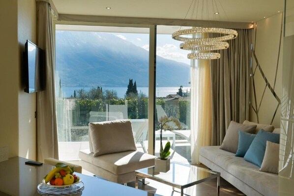 A modern living room with a sofa, a coffee table, and a TV. There is a large window with a view of mountains and a lake.