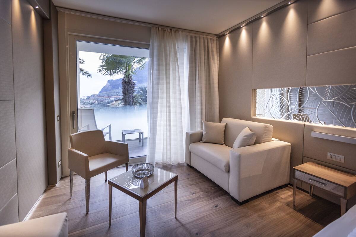 A modern hotel room with a sofa, armchair, and coffee table. There is a large window with a view of a palm tree and mountains.
