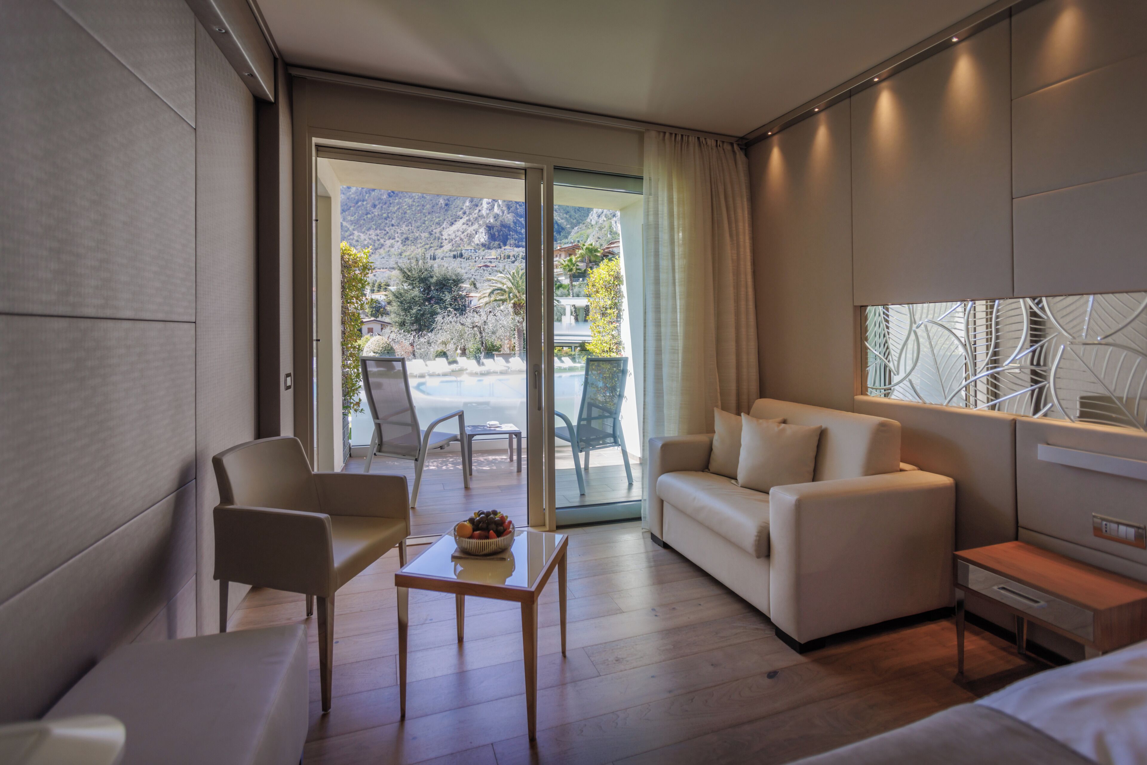 Una camera d'albergo moderna con un divano, un piccolo tavolo con della frutta, una sedia, un letto e vista sulle montagne attraverso una porta in vetro.