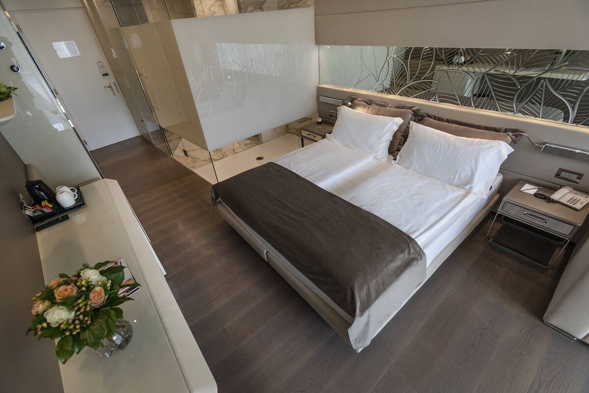 A modern hotel room with a large bed, a bedside table, a mirror, and a view of the outdoors.