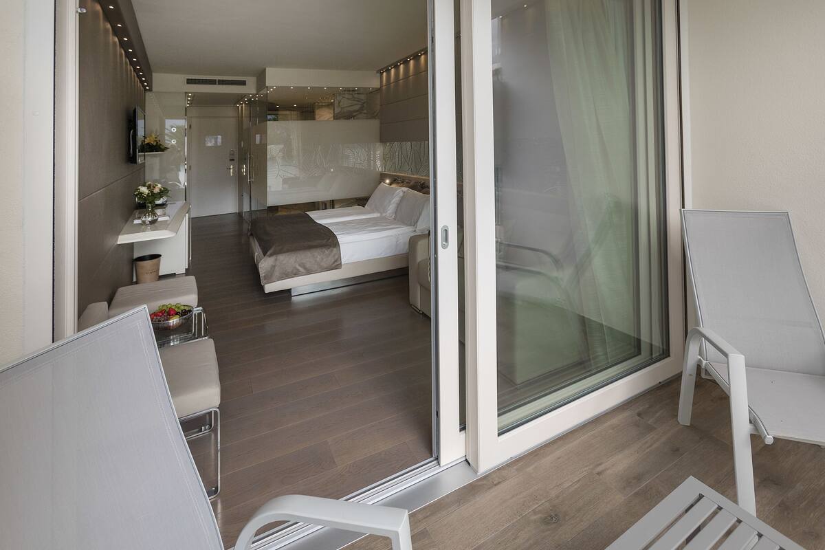 A modern hotel room with a bed, a small sitting area with a table and chairs, a balcony with a glass door, and a bathroom with a shower and toilet.