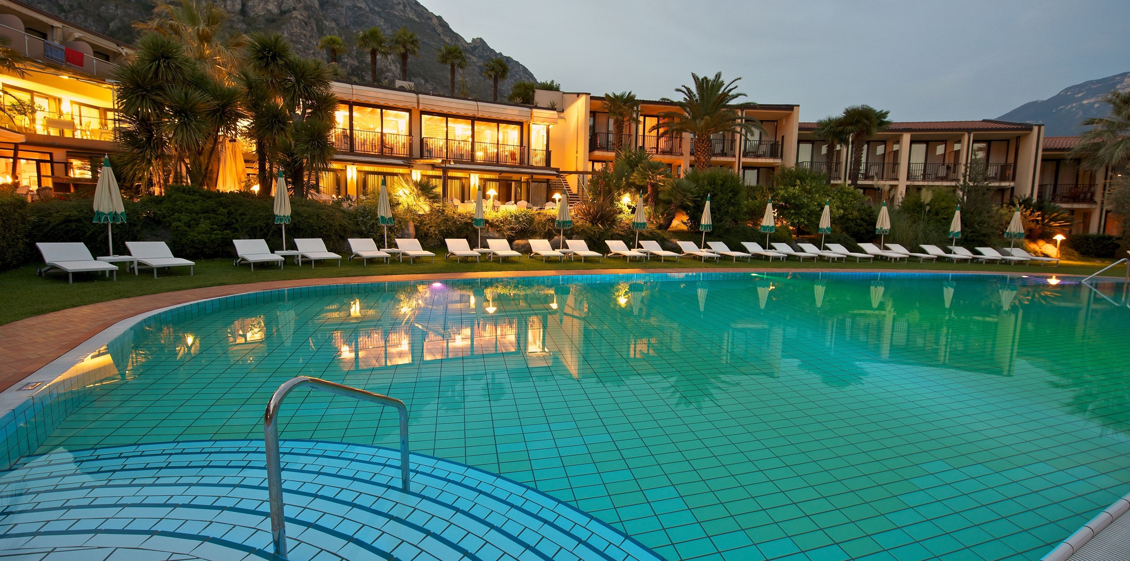 A swimming pool with lounge chairs and umbrellas, surrounded by a building with balconies and palm trees.