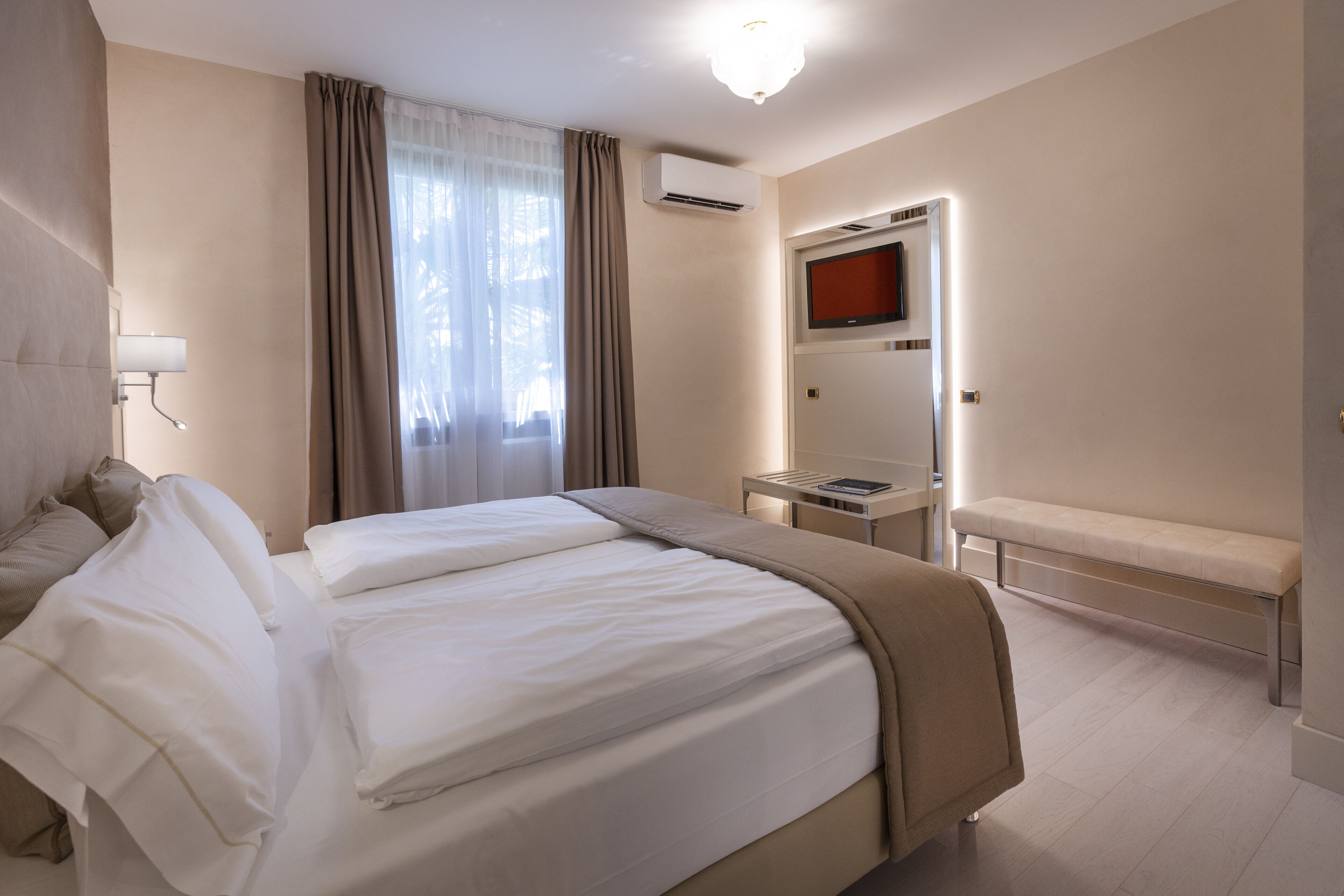 Habitación de hotel con una cama grande, una mesita de noche, un banco, un televisor y un aire acondicionado.