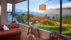 A room with a large window offering a view of a lake and mountains. There is a telescope on a tripod, a wooden stool with a bottle and glasses, and leather sofas. - Hidden Lodge Queenstown (Queenstown)