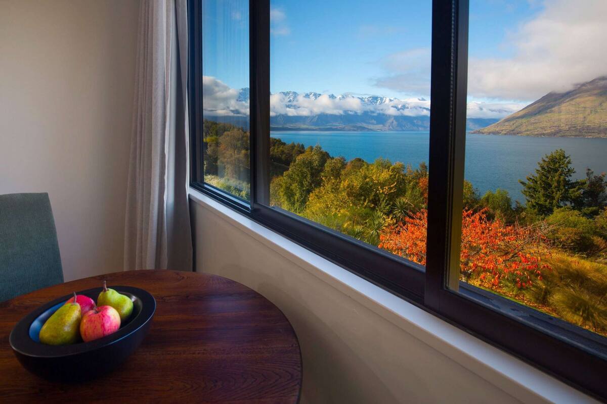 A room with a large window offering a view of a lake, mountains, and autumn foliage.