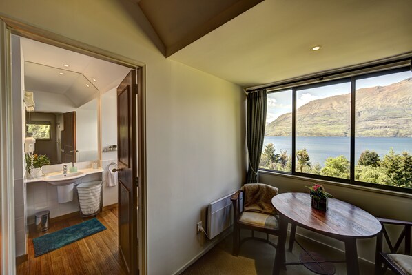 Un baño moderno con lavamanos, espejo y toallero. Una silla y una mesa están ubicadas cerca de un ventanal con vistas a un lago y montañas.