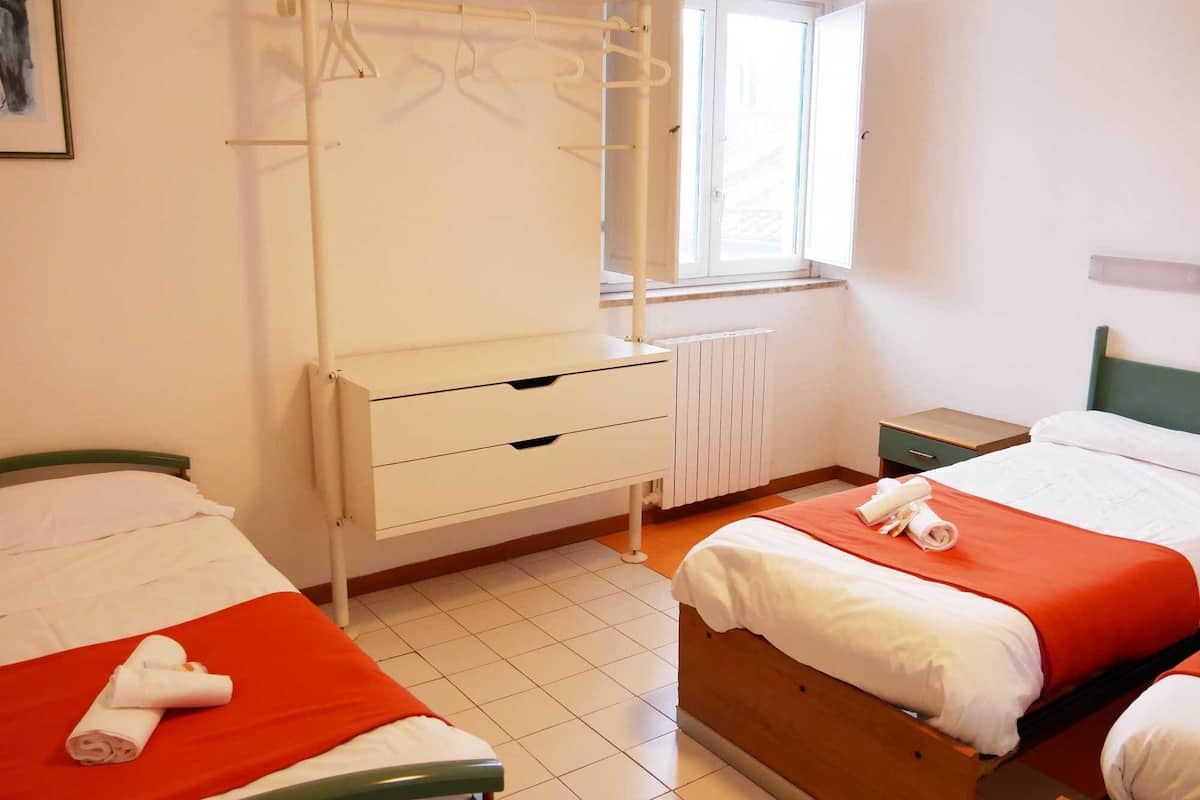 A hotel room with two beds, a window, a radiator, and a wardrobe with hangers.