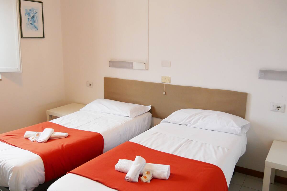 A hotel room with two beds, each with white linens and orange bedspreads. There are white nightstands with lamps, a painting on the wall, and a window with blinds.