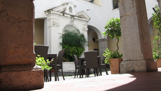 Un patio al aire libre con un edificio blanco con arcos y columnas, rodeado de plantas en macetas y muebles de mimbre.