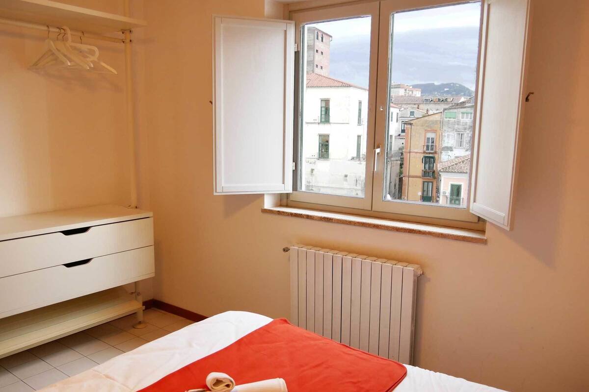 A bedroom with a bed, a radiator, a window with a view of buildings, and a wardrobe with hangers.