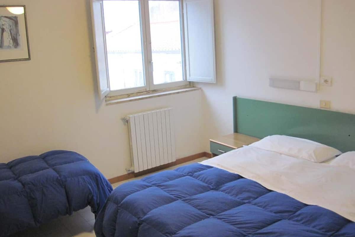 A dormitory room with two beds, a radiator, a window, and a green headboard.