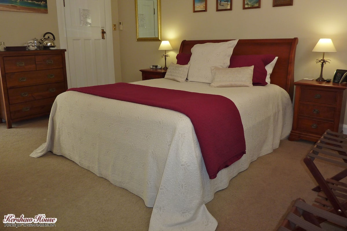 A neatly made bed with a red and white bedspread, two bedside lamps, a wooden nightstand, a wooden dresser, and a door with a white frame.