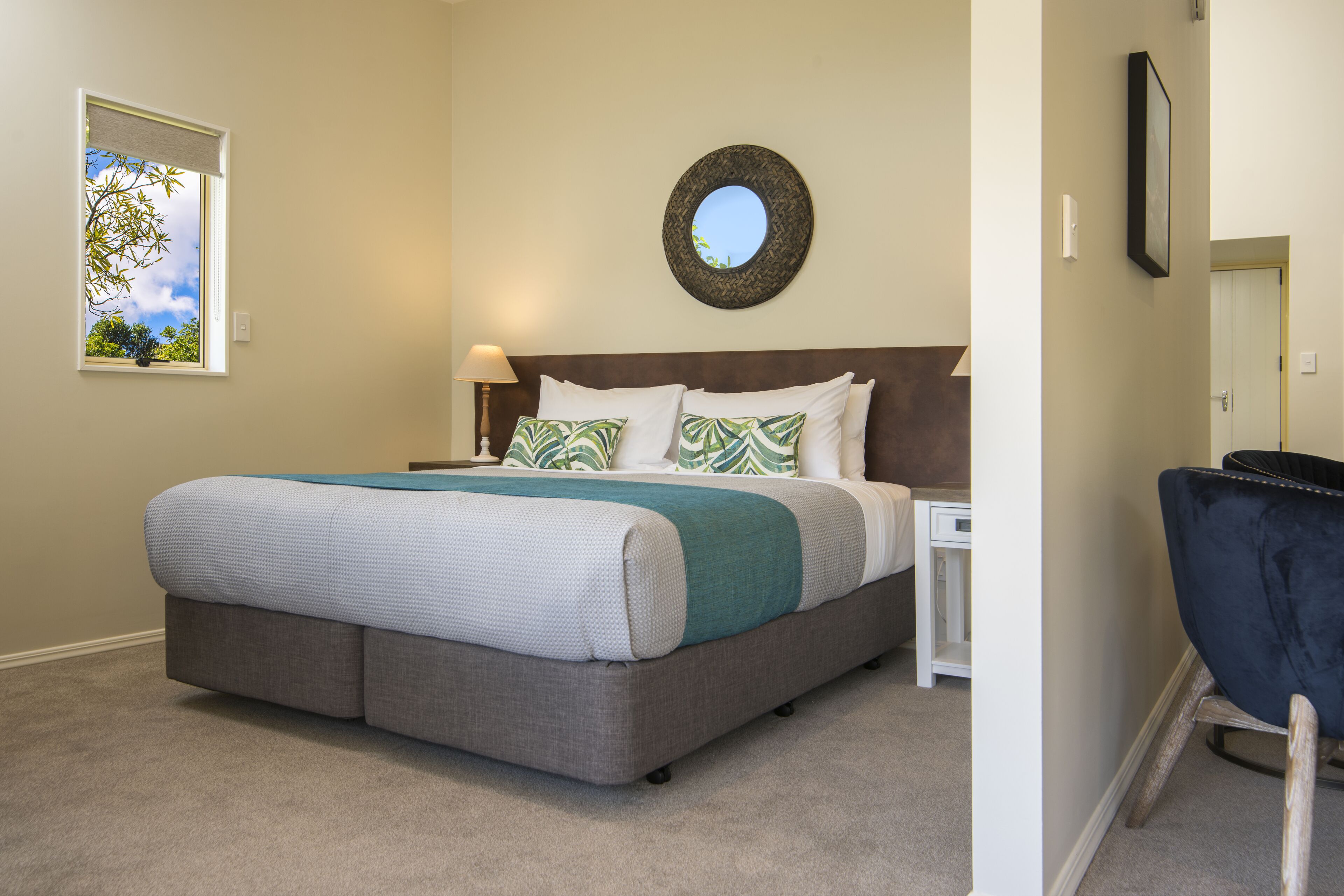 A bedroom with a bed, a nightstand, a chair, a window with a view of trees, and a mirror on the wall.