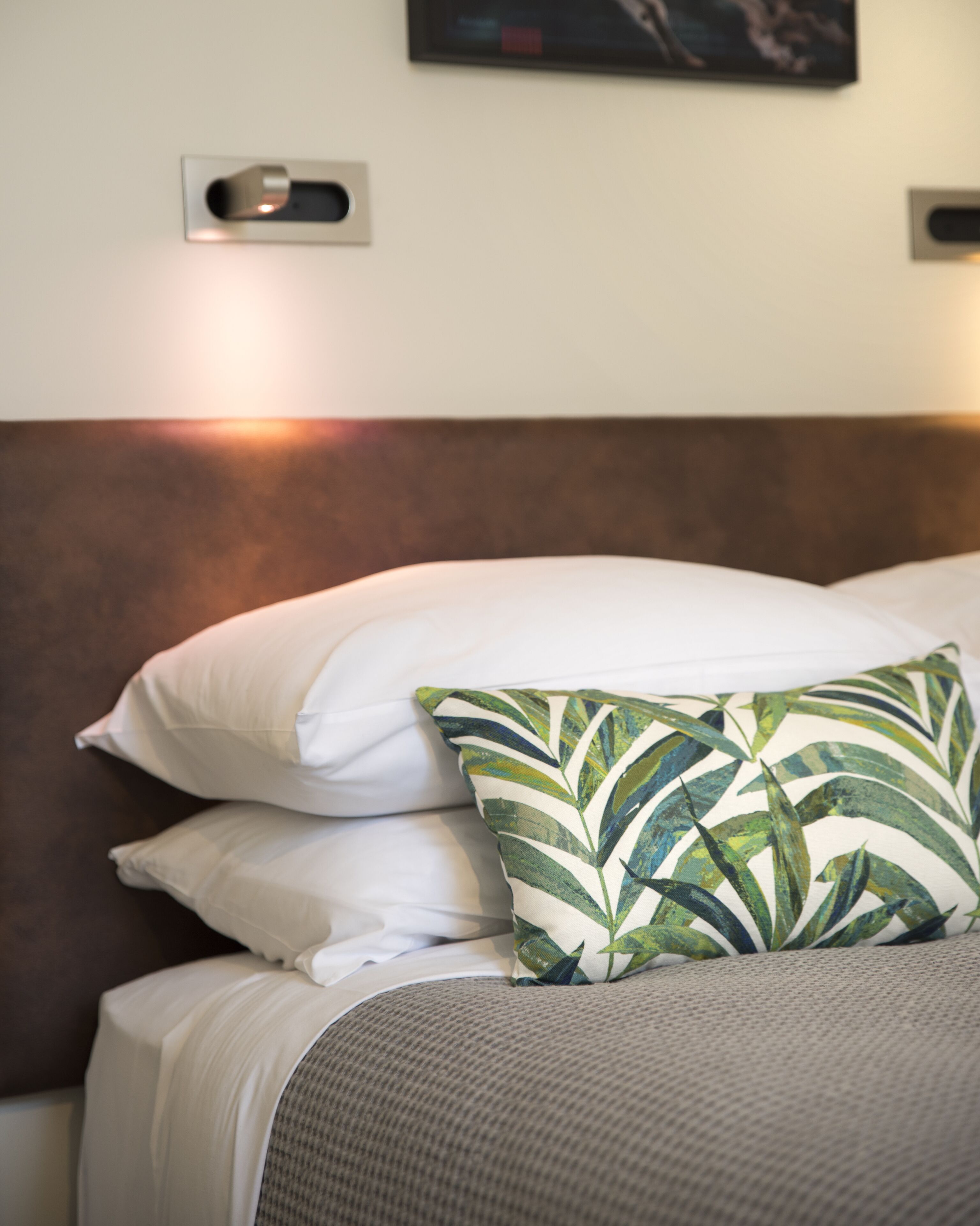 Una cama bien hecha con almohadillas blancas y una almohada estampada con hojas verdes, un cabecero marrón y una lámpara de pared pequeña.