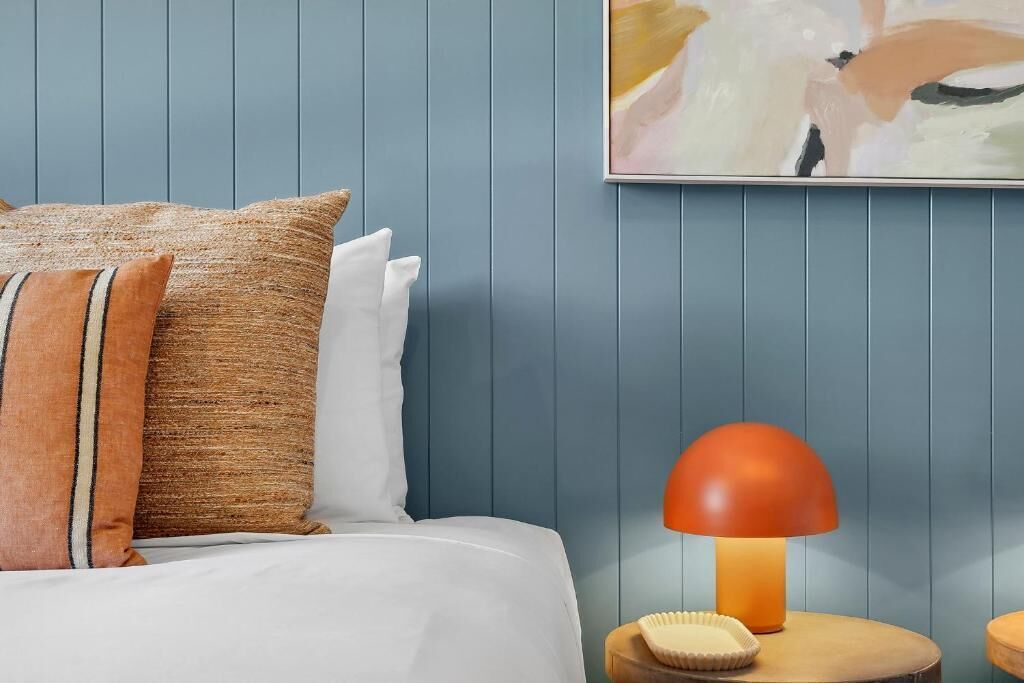 Un lit bien fait, avec une literie blanche, deux oreillers marron et une seule lampe orange posée sur une table de chevet en bois.