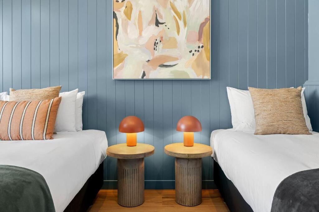 Een hotelkamer met twee bedden, elk met een witte spreidek en meerdere kussens. Tussen de bedden staan twee ronde houten nachtkastjes met lampen. De muren zijn blauw en er hangt een groot abstract schilderij boven de bedden.