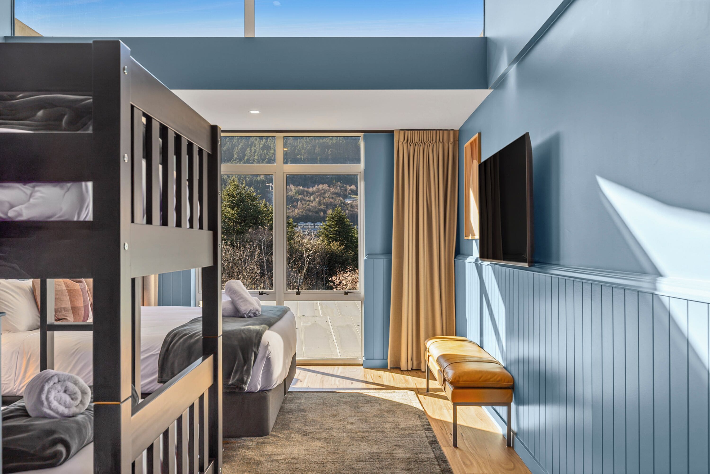 A bedroom with bunk beds, a sofa, a bench, a TV, and a large window with a view of trees and hills.