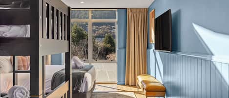 A bedroom with bunk beds, a sofa, a bench, a TV, and a large window with a view of trees and hills.