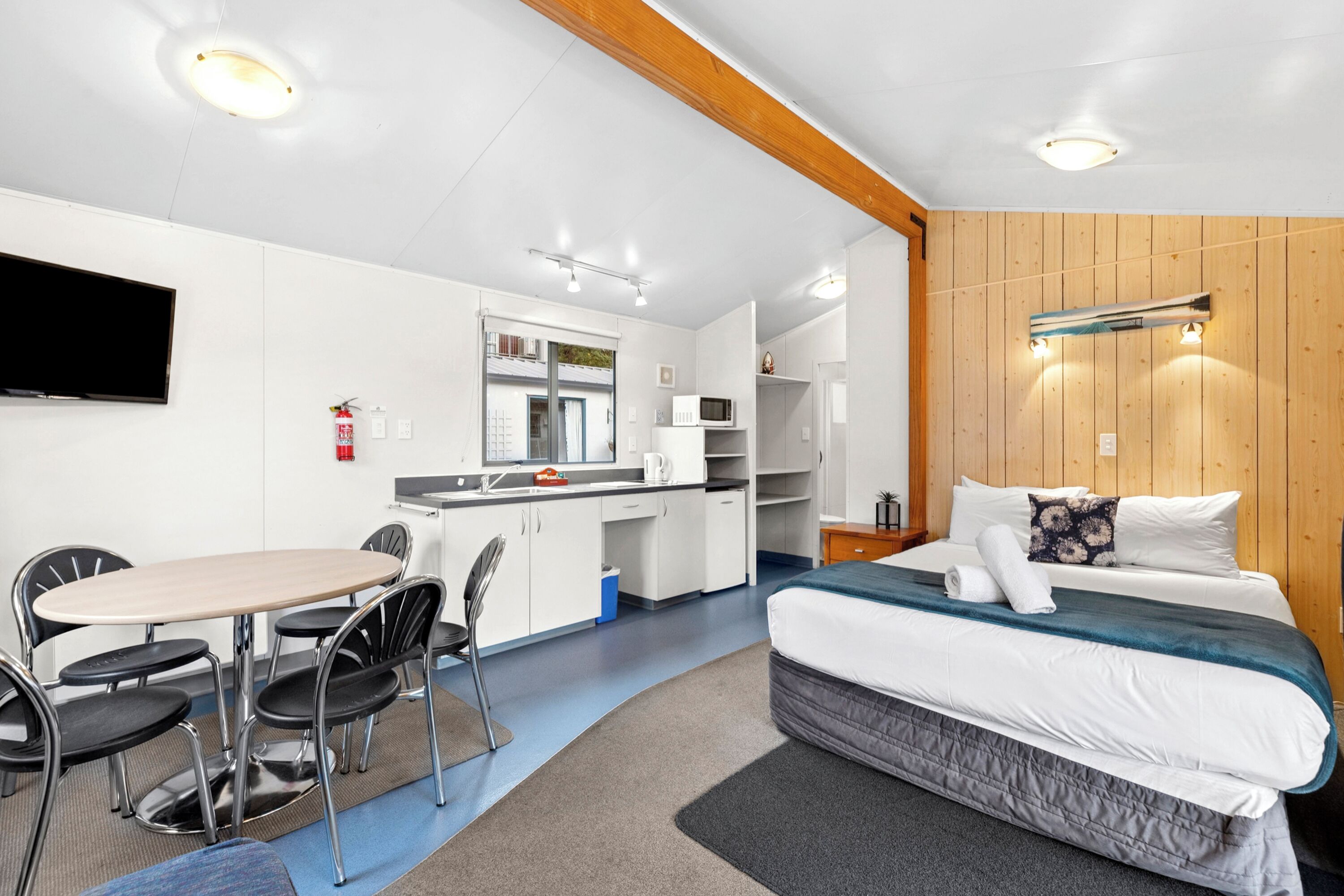 Quarto de hotel compacto com cama, pequeno espaço de cozinha, mesa de jantar com cadeiras e TV fixada na parede.