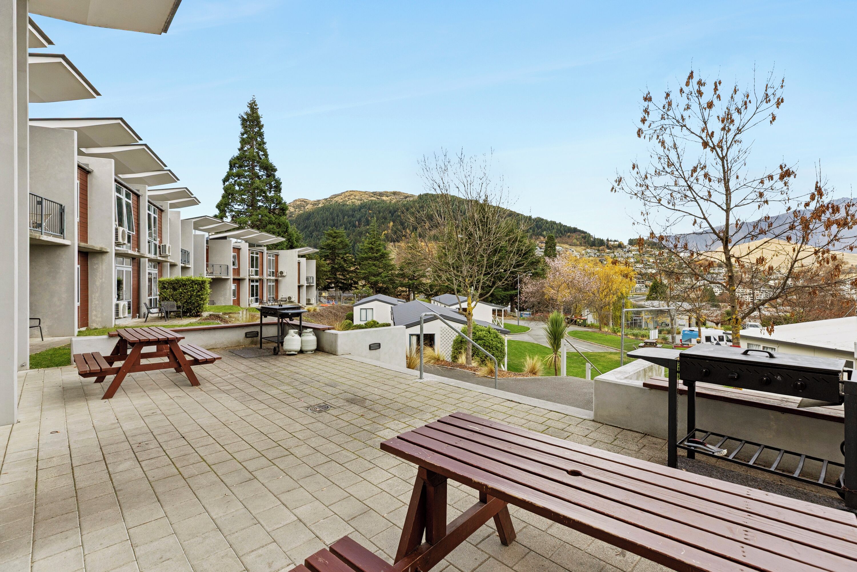 A patio area with picnic tables and a barbecue grill, overlooking a residential neighborhood and hills.