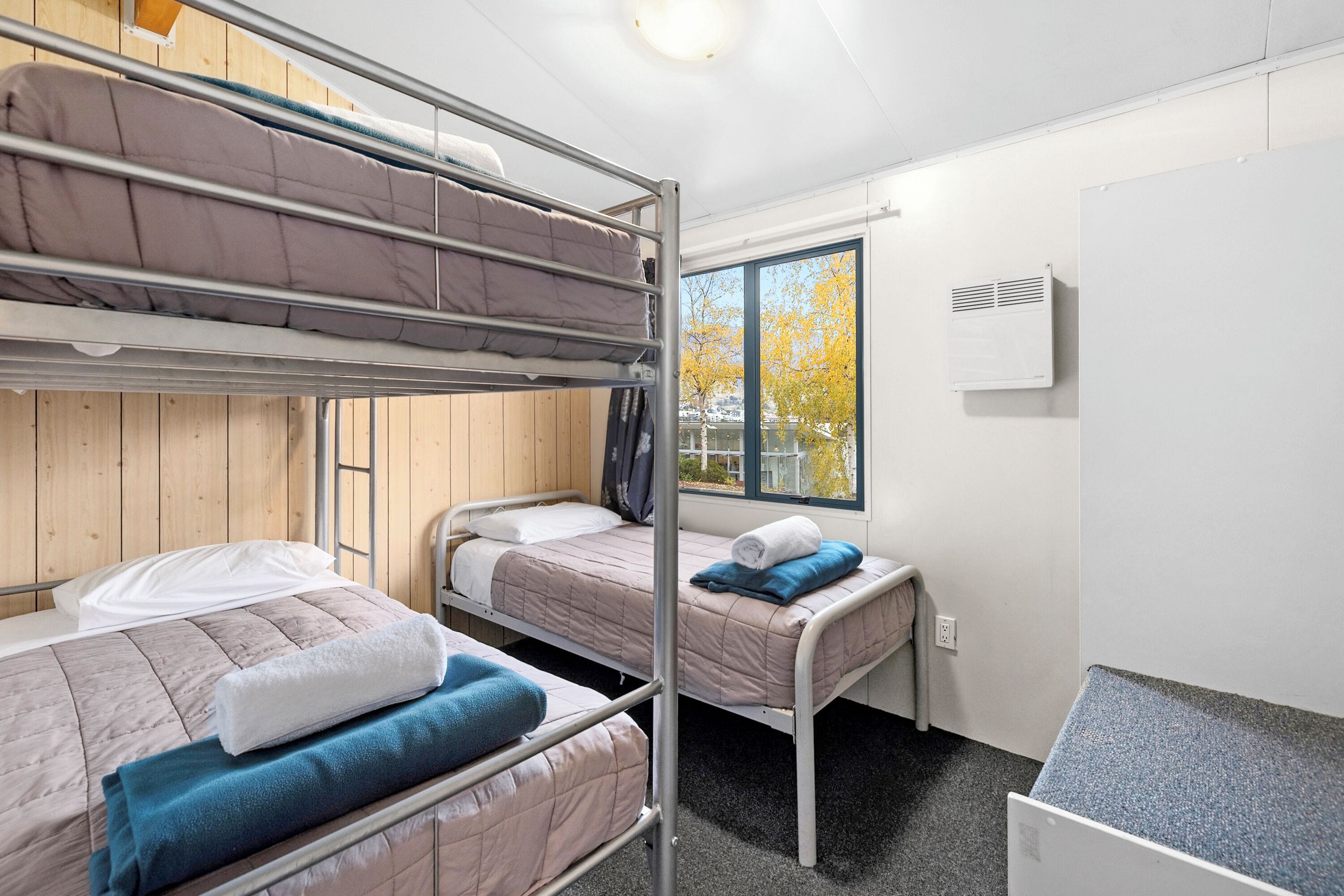 A dormitory room with bunk beds, a window with a view of trees, and an air conditioning unit on the wall.