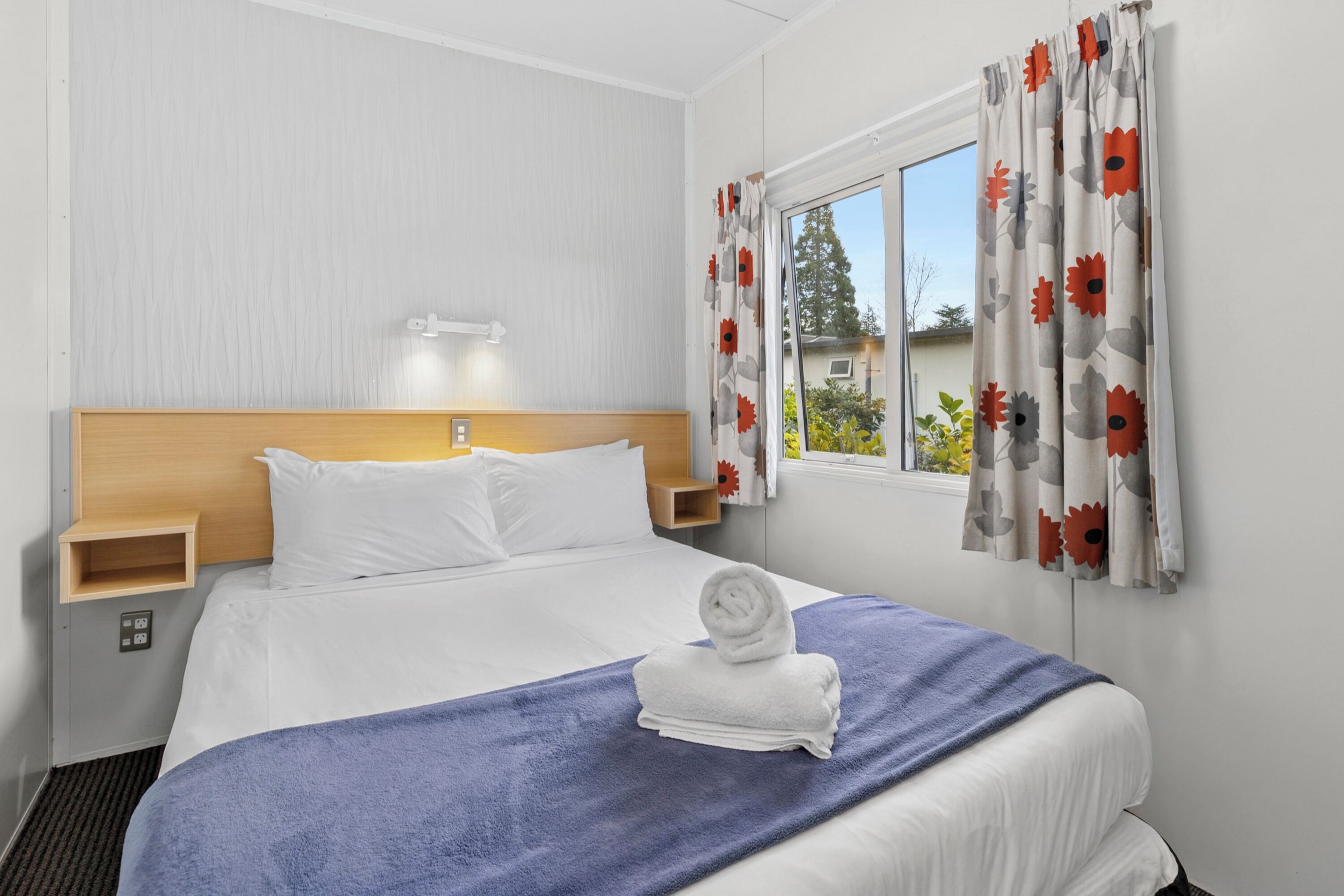 A hotel room with a bed, a nightstand, a window with curtains, and a view of the outdoors.