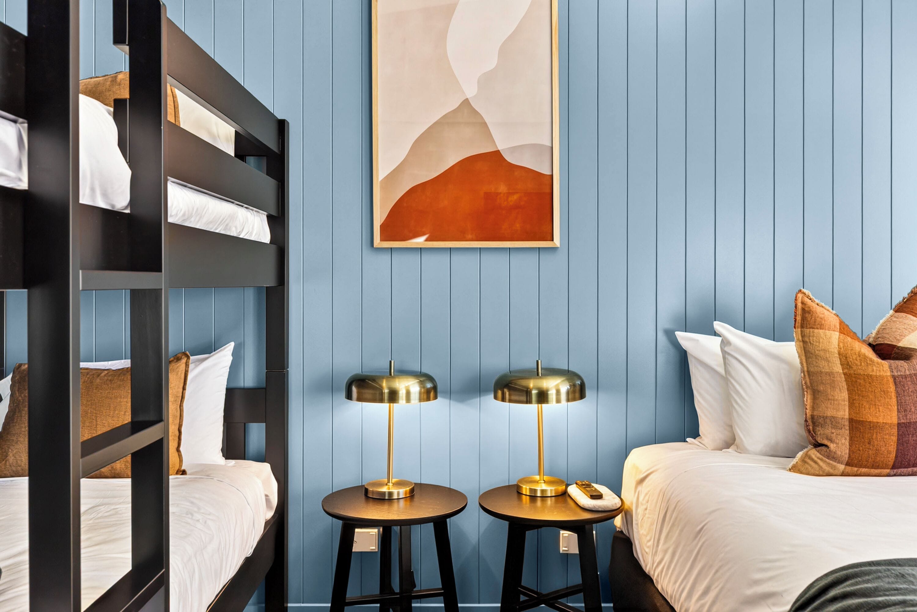 A room with bunk beds, two bedside tables with lamps, a painting on the wall, and a bed with pillows.