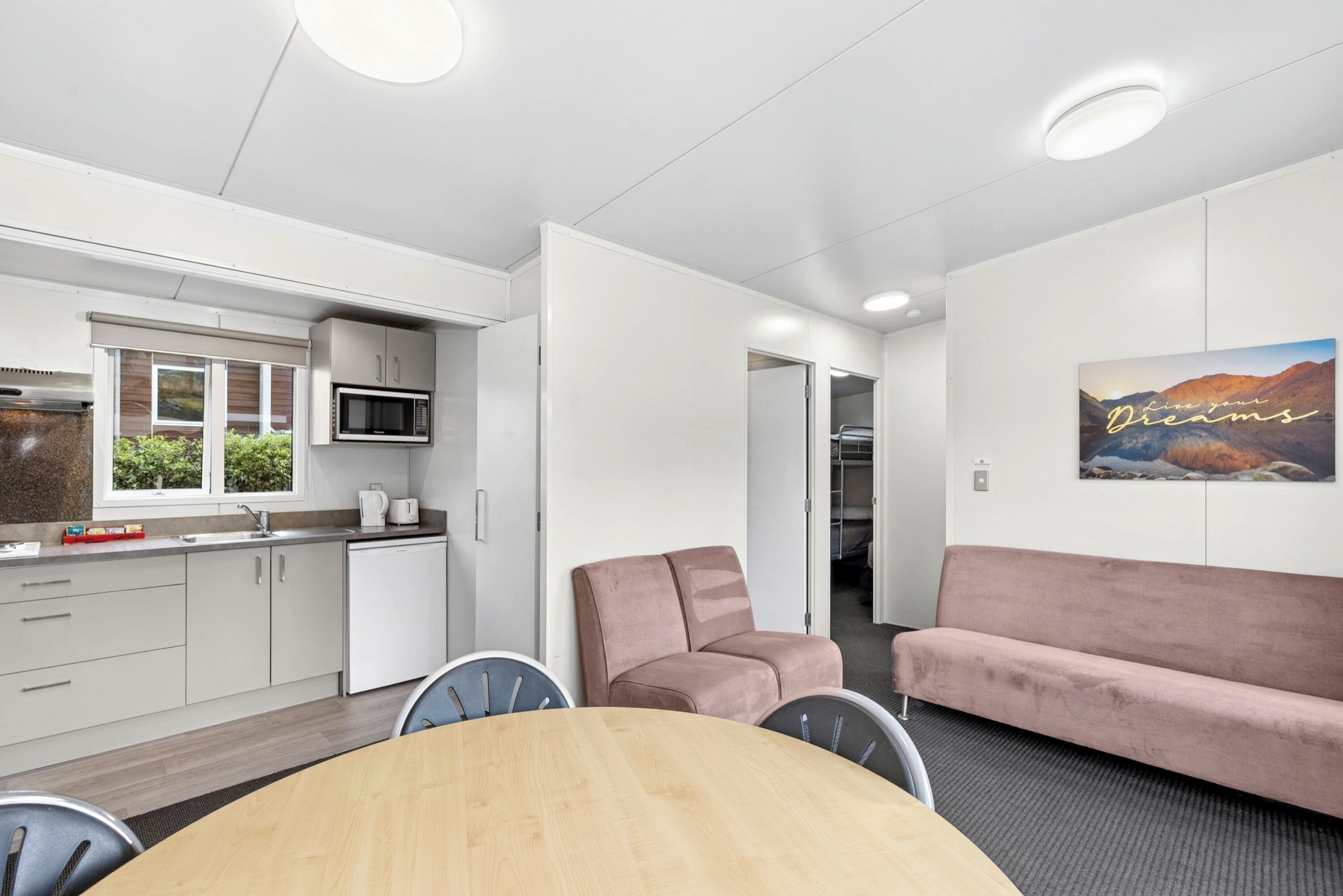 Ett modernt kök med vita skåp, en mikrovågsugn och ett fönster med utsikt över grönområden. Det finns en matplats med ett träbord och stolar, samt en rosa soffa.