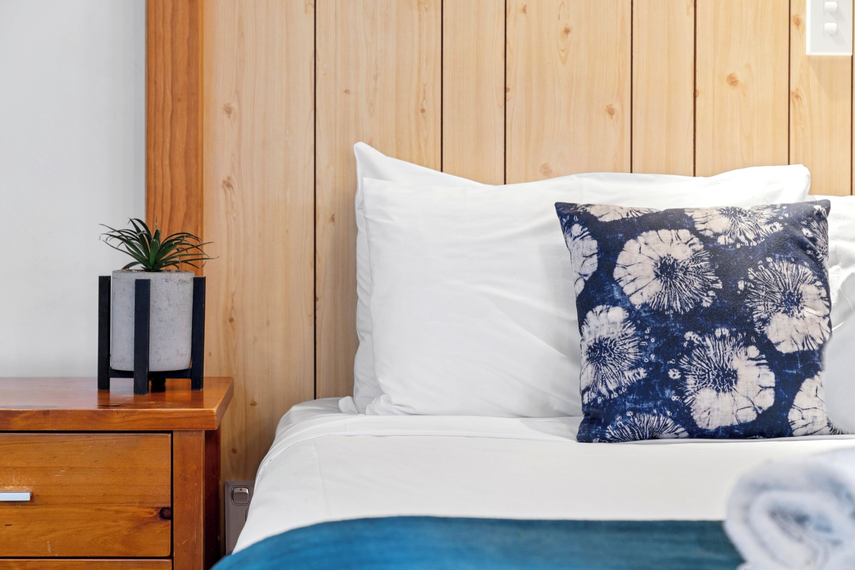 Una cama bien tendida con almohadas blancas y estampadas, una pequeña planta en maceta sobre una mesita de noche de madera, y un panel de madera en la pared.