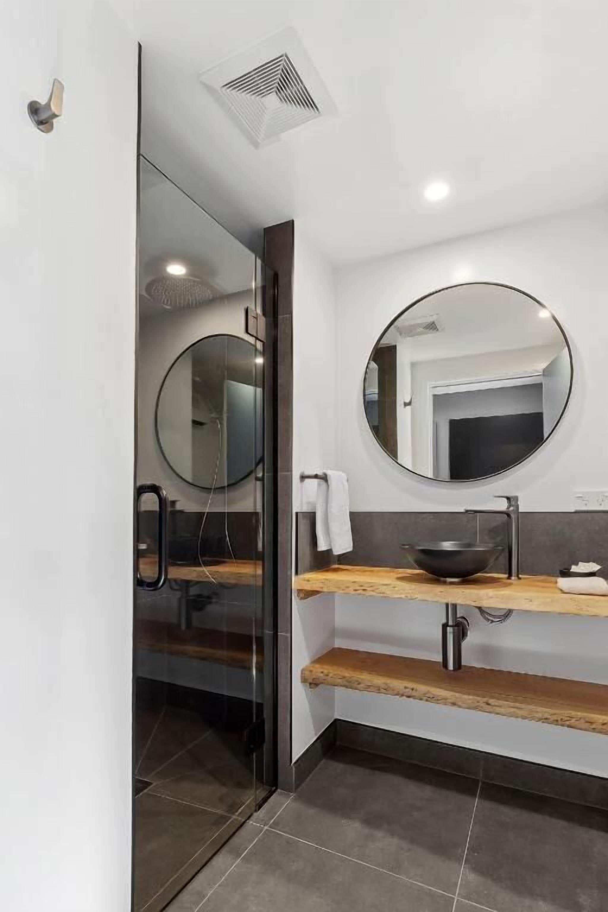 A modern bathroom with a glass shower enclosure, a round mirror, and a wooden vanity with a sink.
