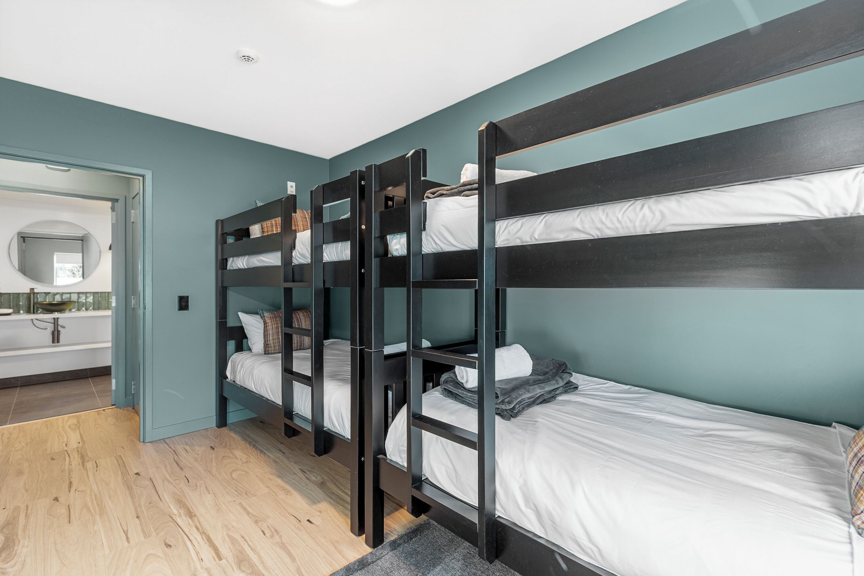 A room with bunk beds, a bathroom with a mirror, and a wooden floor.