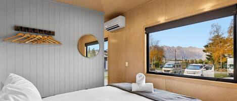 Una habitación de hotel con cama, ventana con vistas a la montaña, espejo y unidad de aire acondicionado.